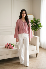 Front view of a woman wearing a tie-front blouse with long bell sleeves, paired with white high-waisted wide-leg trousers. The blouse features two delicate bow closures at the chest.