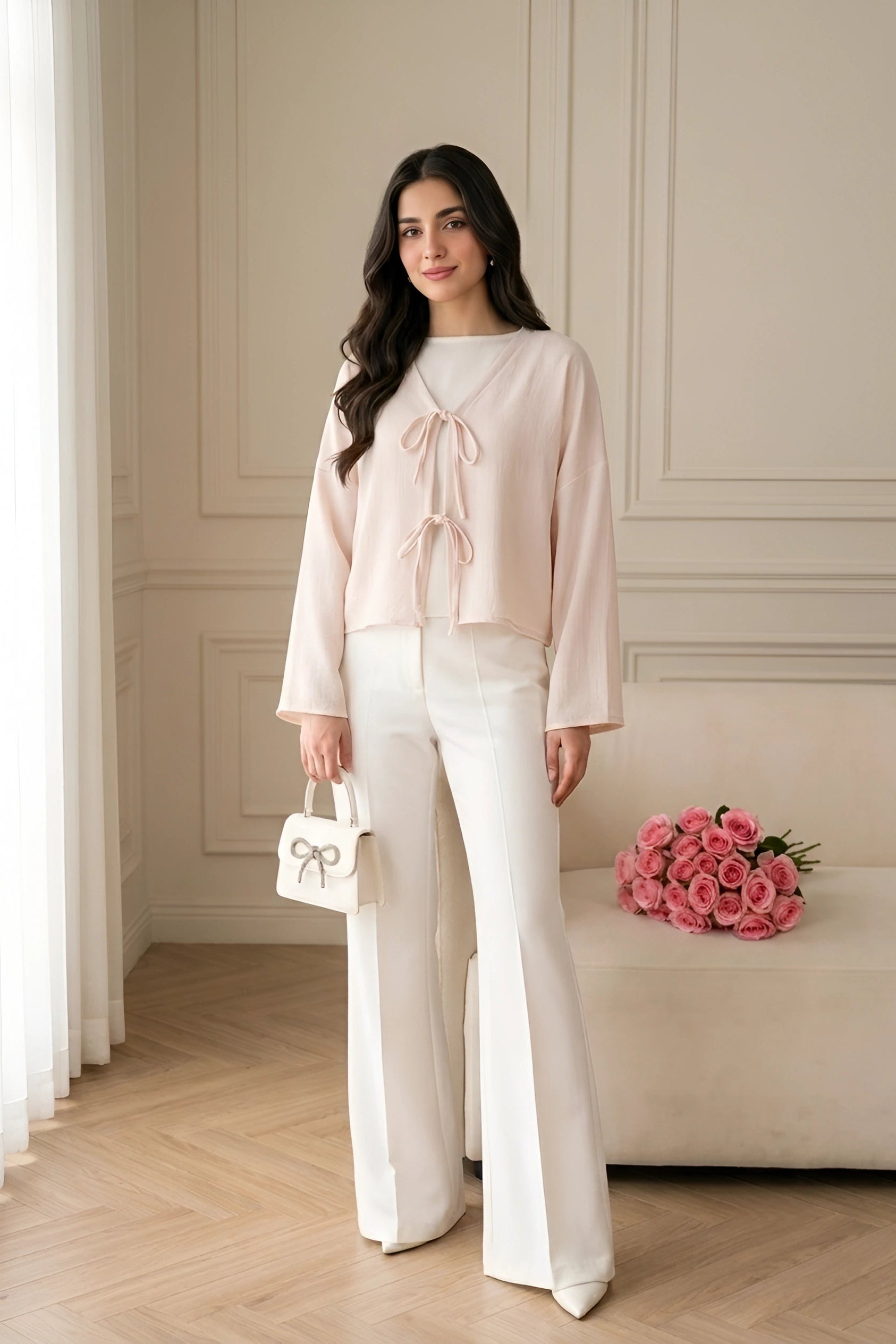 A woman models a light pink long-sleeve blouse with a tie-neck detail and white wide-leg trousers, holding a small white handbag with a bow, in a bright, elegant room with a bench of pink roses.