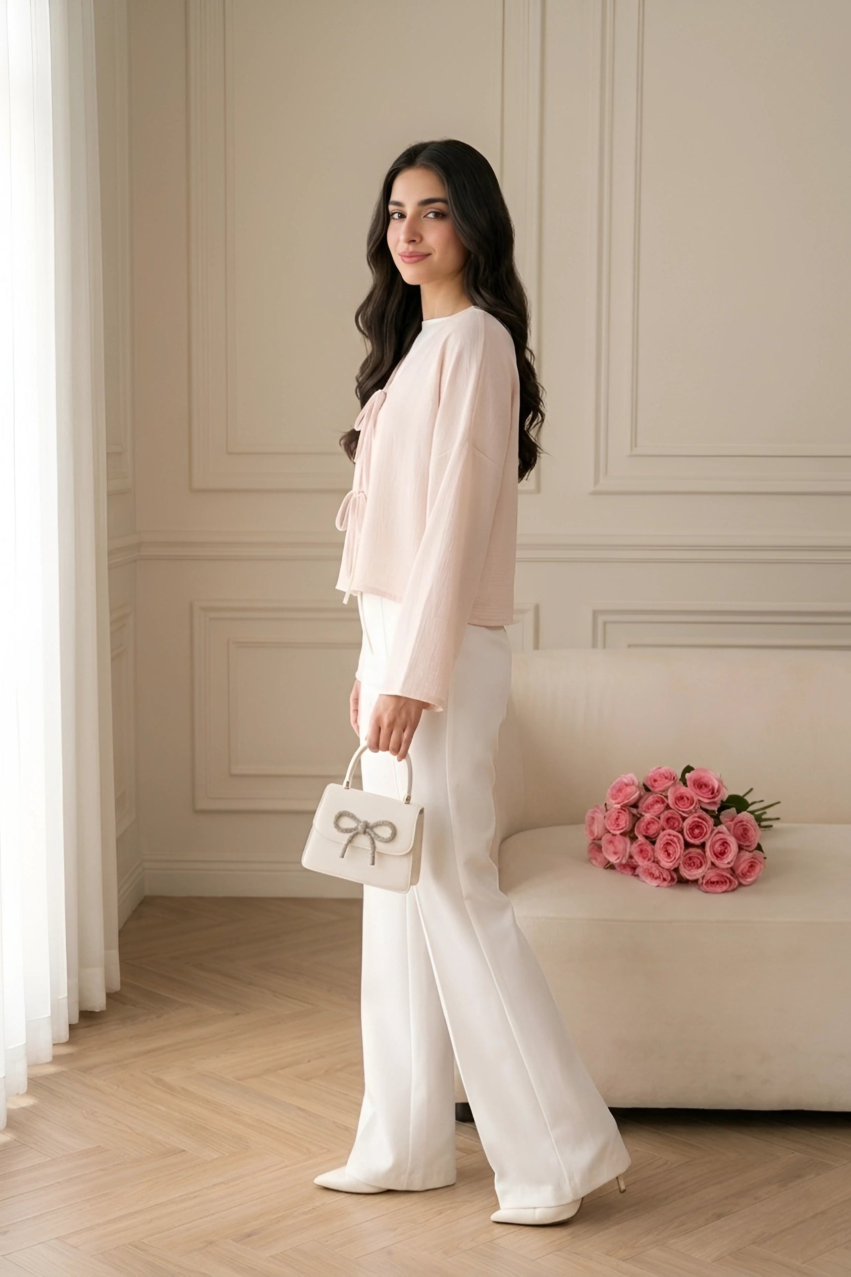 A woman stands in a bright, elegant room wearing a light pink long-sleeve blouse and white wide-leg trousers, holding a small white handbag with a bow detail. A beige sofa with pink roses is in the background.