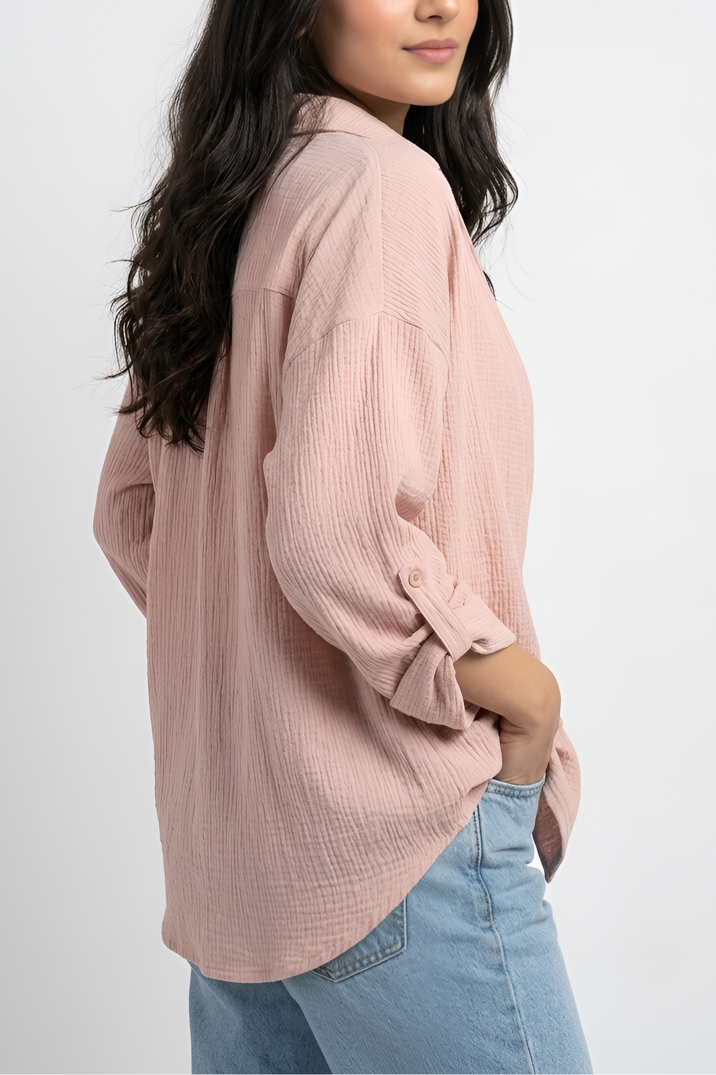 A woman wearing a dusty pink textured long-sleeve blouse and light blue jeans, photographed from the side against a plain light gray studio background.