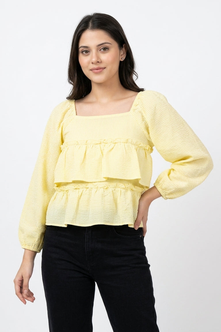 A woman modeling a pale yellow textured long-sleeve blouse with a square neckline and ruffled layers, paired with black pants, against a plain white studio background.