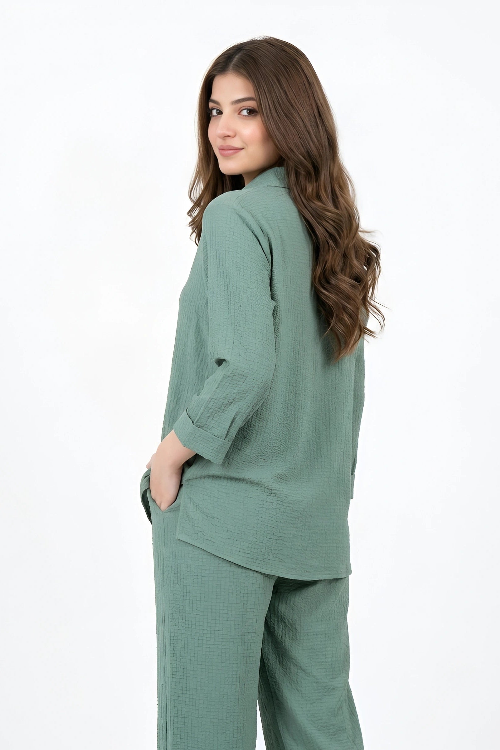 A woman wearing a matching sage green two-piece set consisting of a long-sleeve button-up shirt and wide-leg trousers, shown from a three-quarter back view against a plain white studio background.