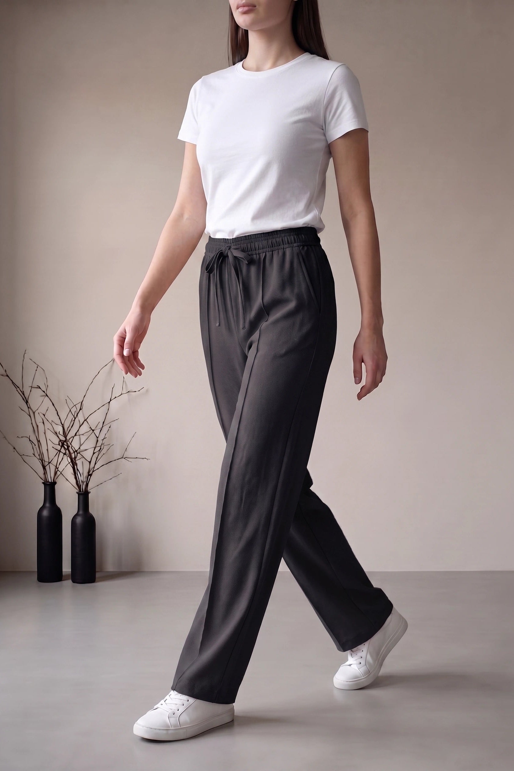 A woman wearing dark charcoal gray drawstring wide-leg pants, a white t-shirt, and white sneakers, walking in a minimalist studio setting with neutral beige walls and two black vases with dried branches.