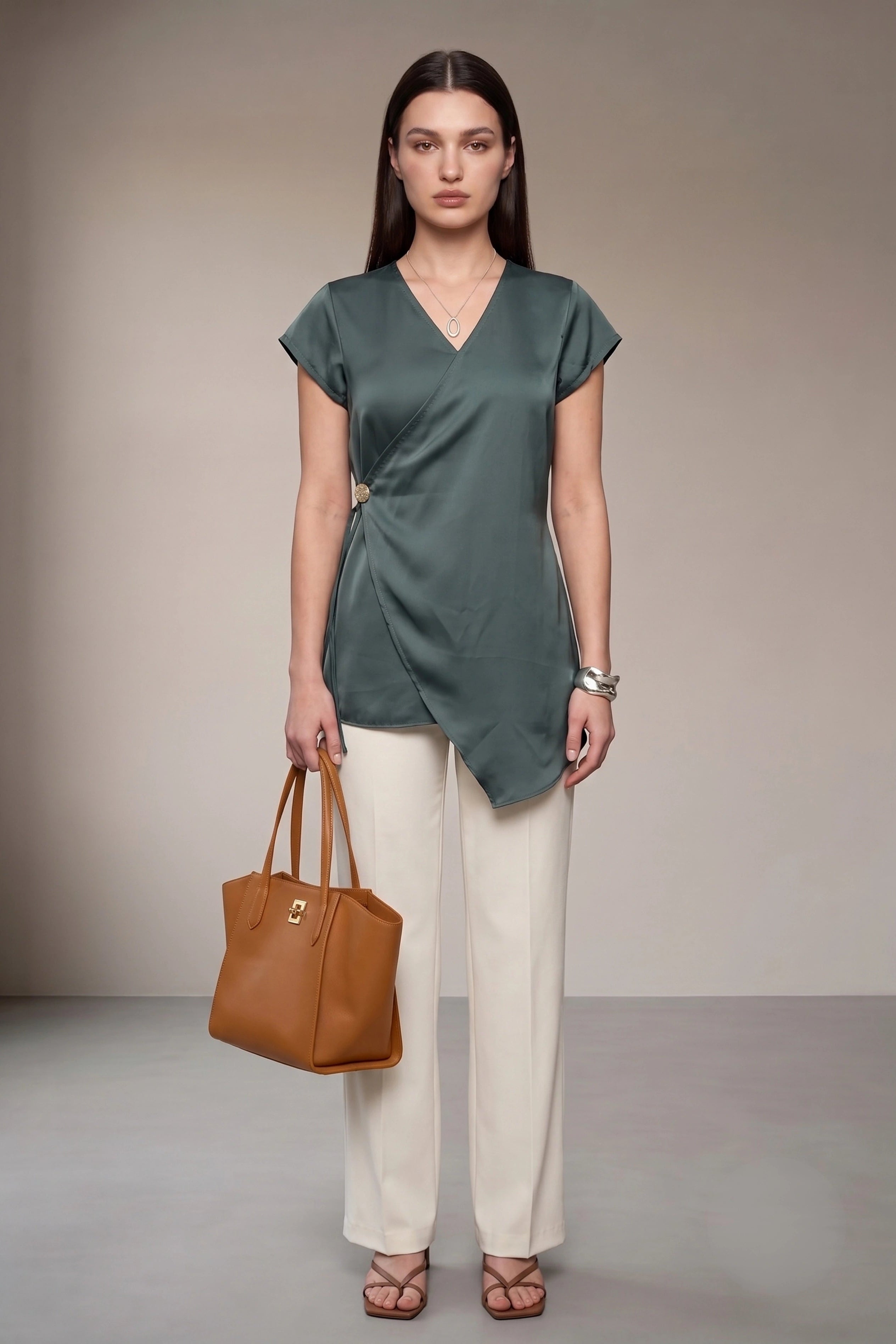 A woman stands in a studio wearing a slate grey satin wrap top with cream wide-leg trousers, holding a brown leather tote bag.