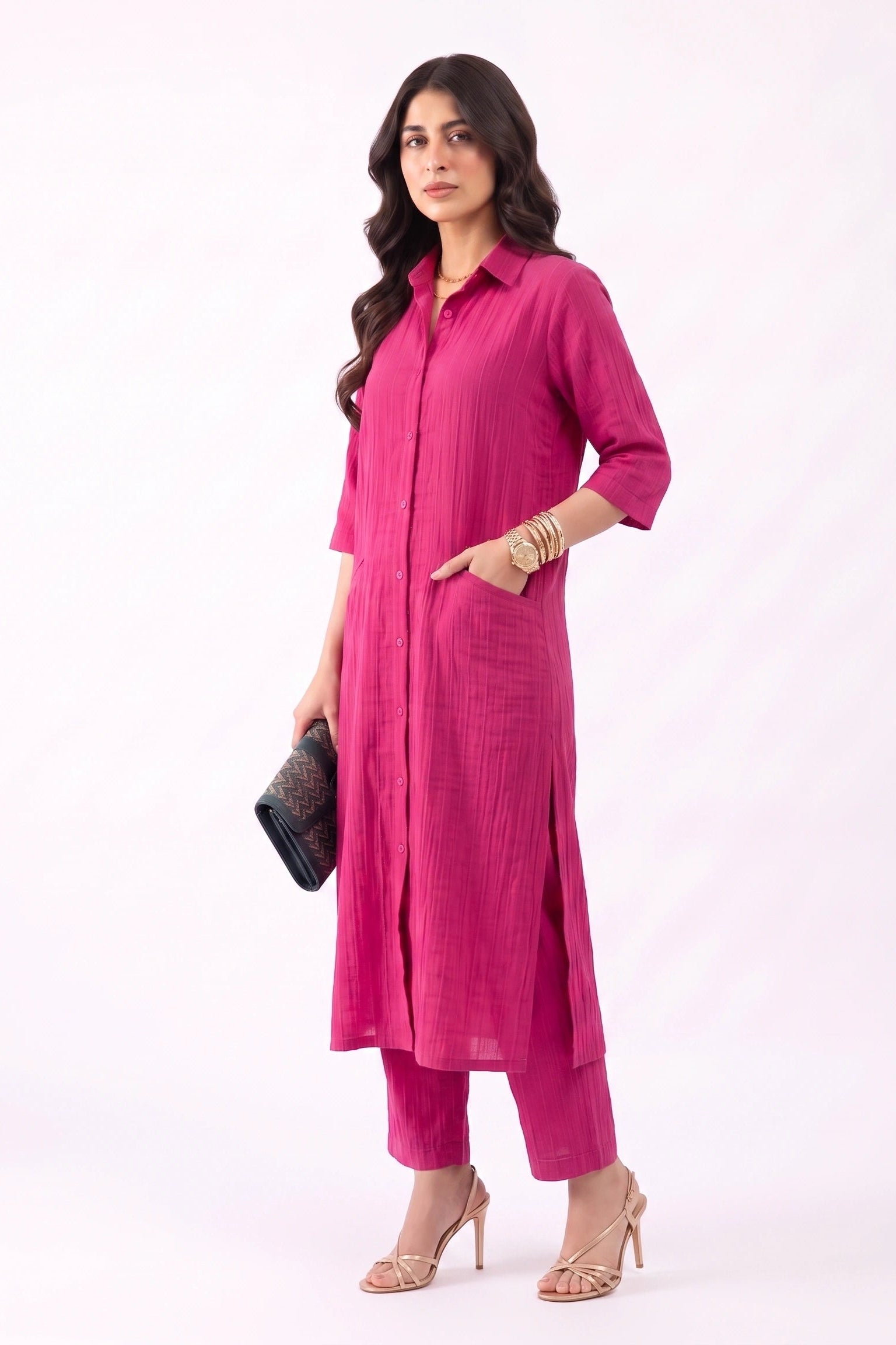 A woman modeling a bright pink kurta set with matching pants, holding a black clutch purse, against a plain white studio background.