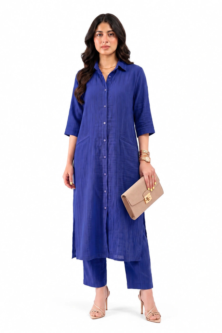 A woman modeling a royal blue button-up kurta with matching cropped pants, holding a beige clutch purse against a plain white background.
