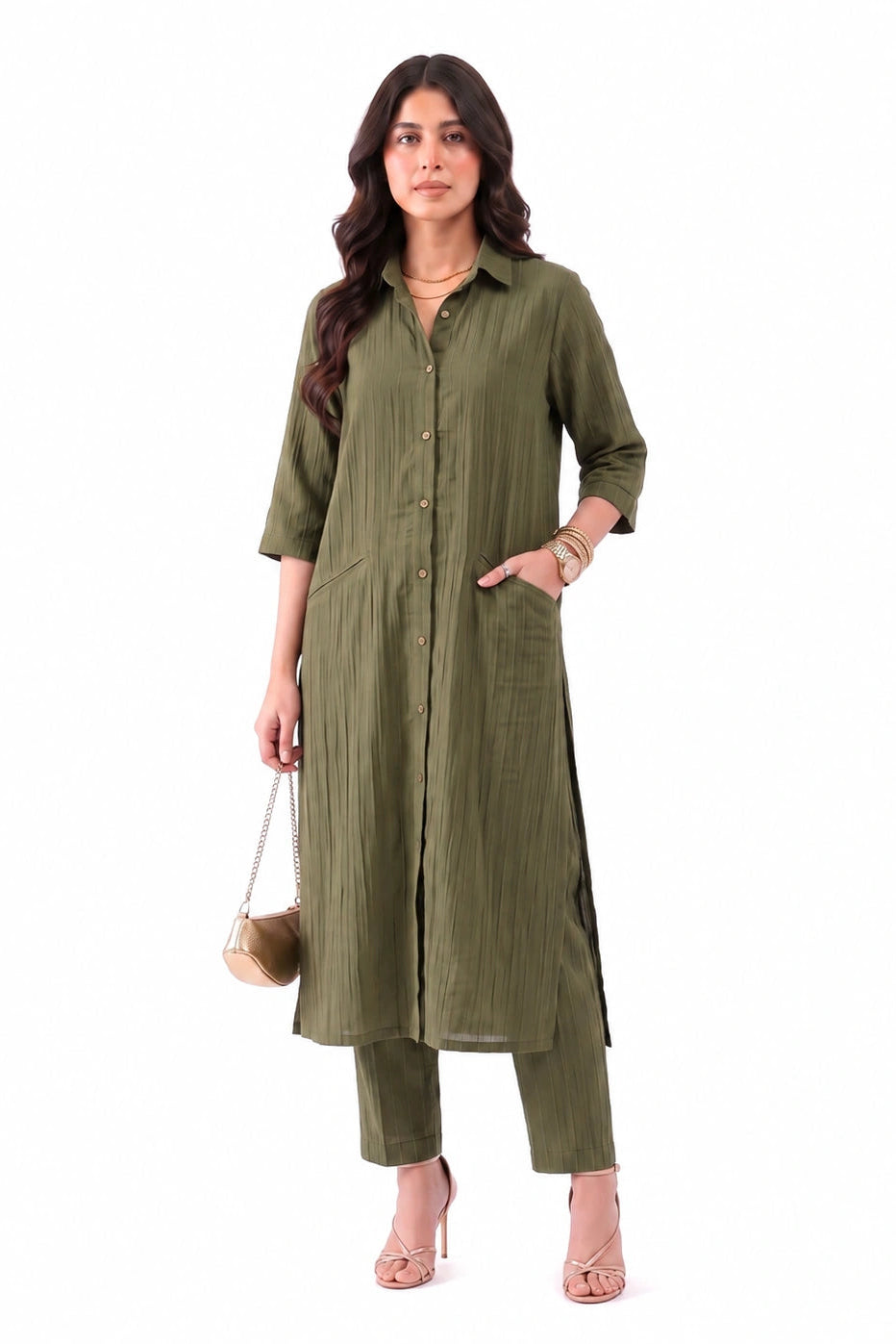 A woman modeling an olive green long kurta with matching pants, standing against a plain white background. She is holding a small beige handbag and wearing nude high heels.