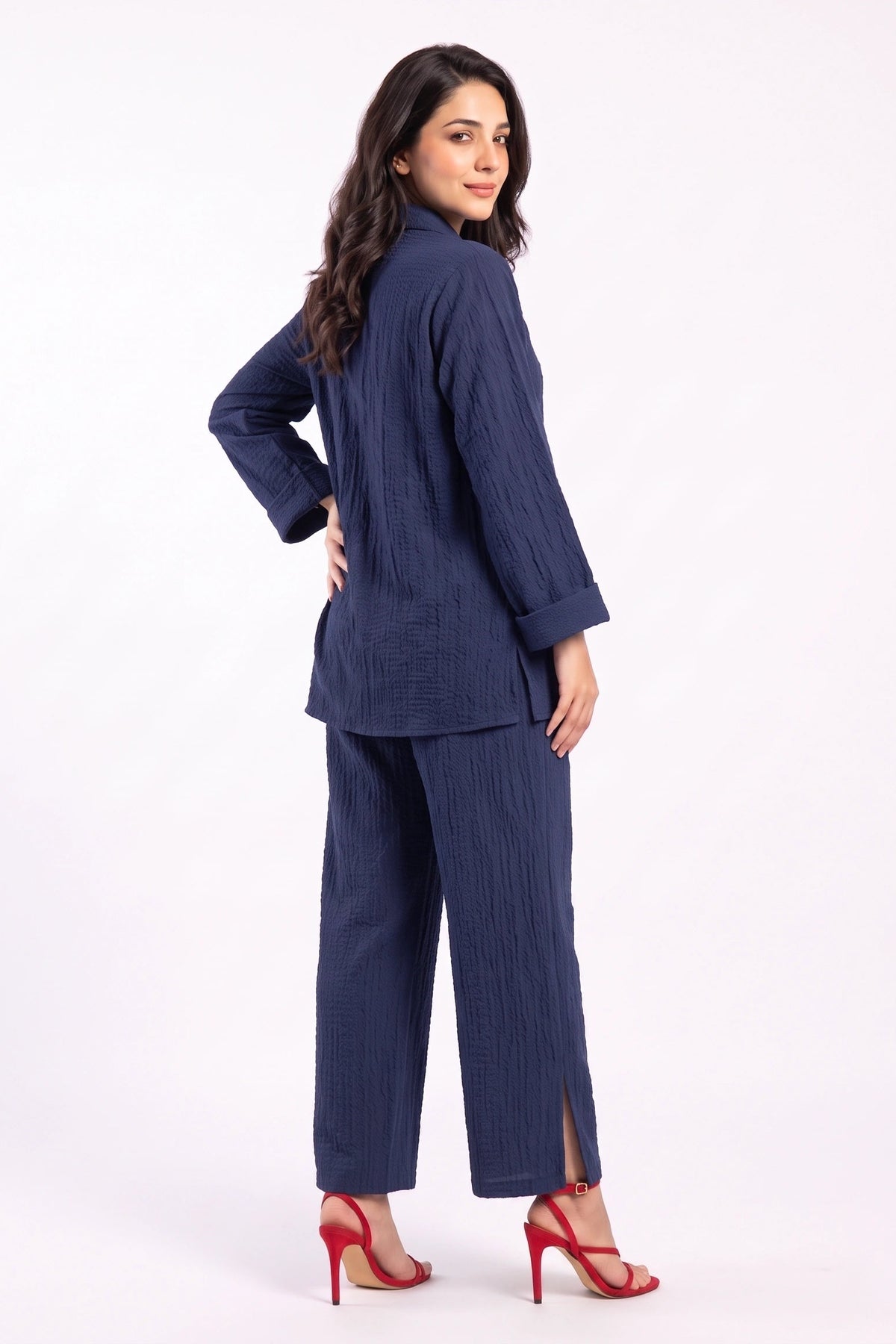 A woman modeling a navy blue two-piece set consisting of a textured long-sleeve top and matching wide-leg trousers, paired with red high-heeled sandals, against a plain white studio background.