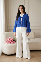 A front-view shot of a model wearing a royal blue blouse featuring double bow-tie front closures and long bell sleeves. The vibrant blue top is styled with white wide-leg trousers and a white handbag, creating a sophisticated and airy aesthetic.
