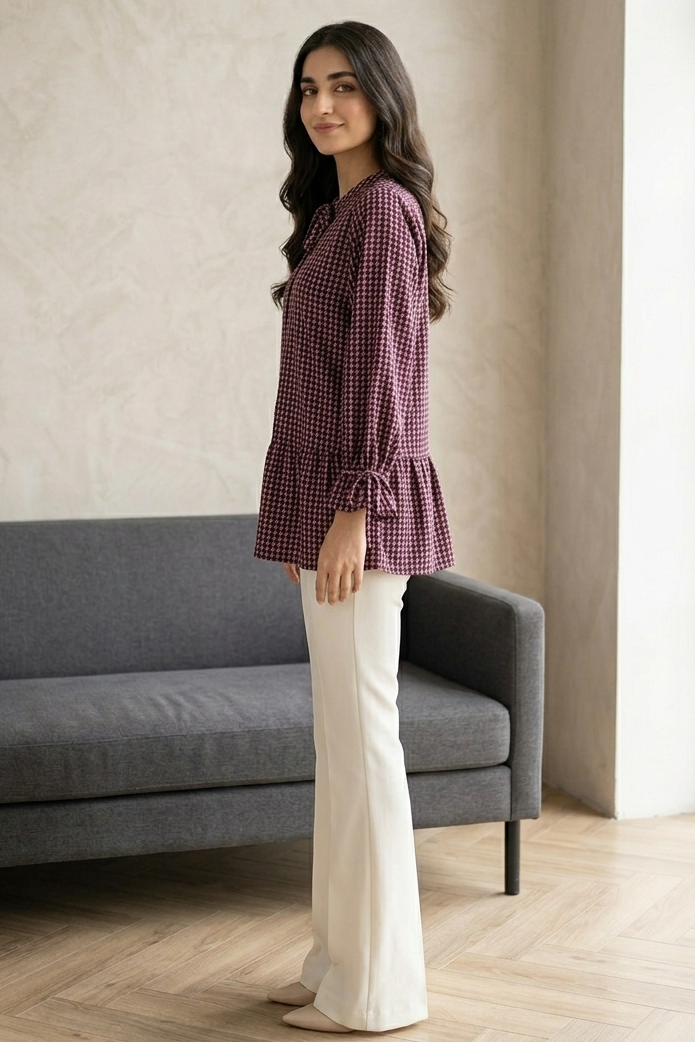 Woman in a purple checkered blouse and white pants standing in a room with a gray sofa and large window.