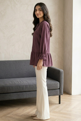 Woman in a purple checkered blouse and white pants standing in a room with a gray sofa and large window.