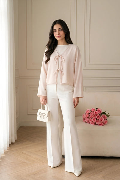 A woman models a light pink long-sleeve blouse with a tie-neck detail and white wide-leg trousers, holding a small white handbag with a bow, in a bright, elegant room with a bench of pink roses.
