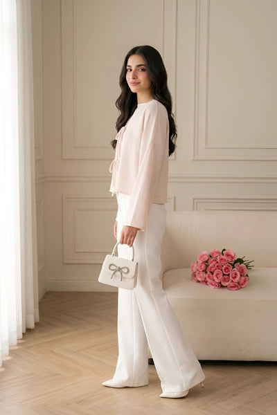 A woman stands in a bright, elegant room wearing a light pink long-sleeve blouse and white wide-leg trousers, holding a small white handbag with a bow detail. A beige sofa with pink roses is in the background.