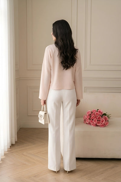 A woman stands with her back to the camera in a bright, elegant room, wearing a light pink long-sleeve top and white wide-leg trousers, holding a small white handbag.