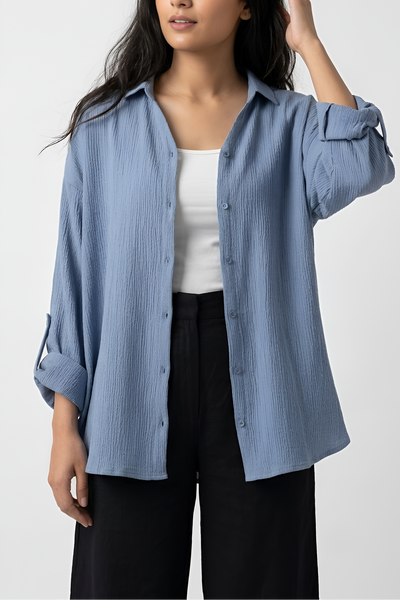A woman modeling a light blue, long-sleeve button-up shirt with a subtle vertical crinkle texture, worn open over a white tank top and black trousers, against a plain light gray background.