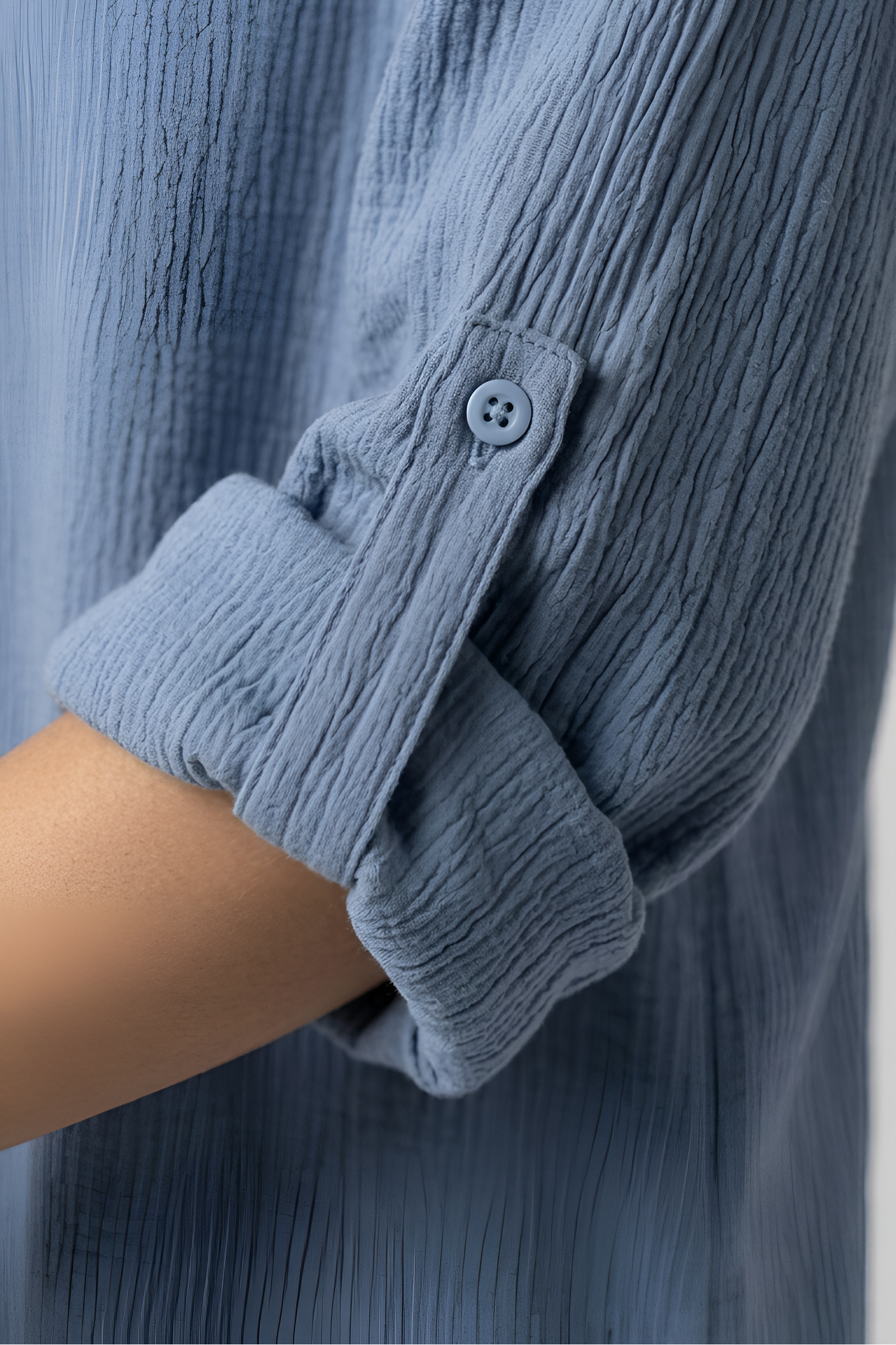 Close-up of a person wearing a crinkled gauze long-sleeve shirt in dusty blue, showing the rolled cuff and button detail.