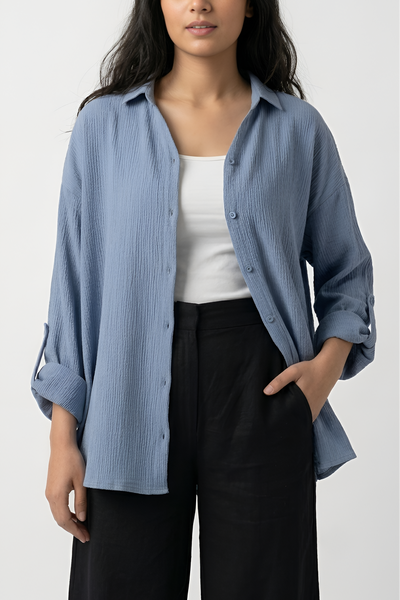 A woman modeling a light blue crinkle-textured button-up shirt over a white tank top and black wide-leg pants, photographed against a plain light gray studio background.