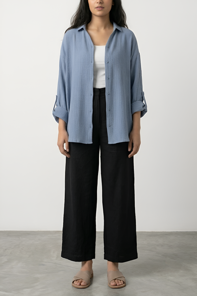 A woman stands in a studio wearing a light blue button-up shirt with rolled sleeves, a white tank top, black wide-leg cropped pants, and tan slide sandals against a plain light gray background.