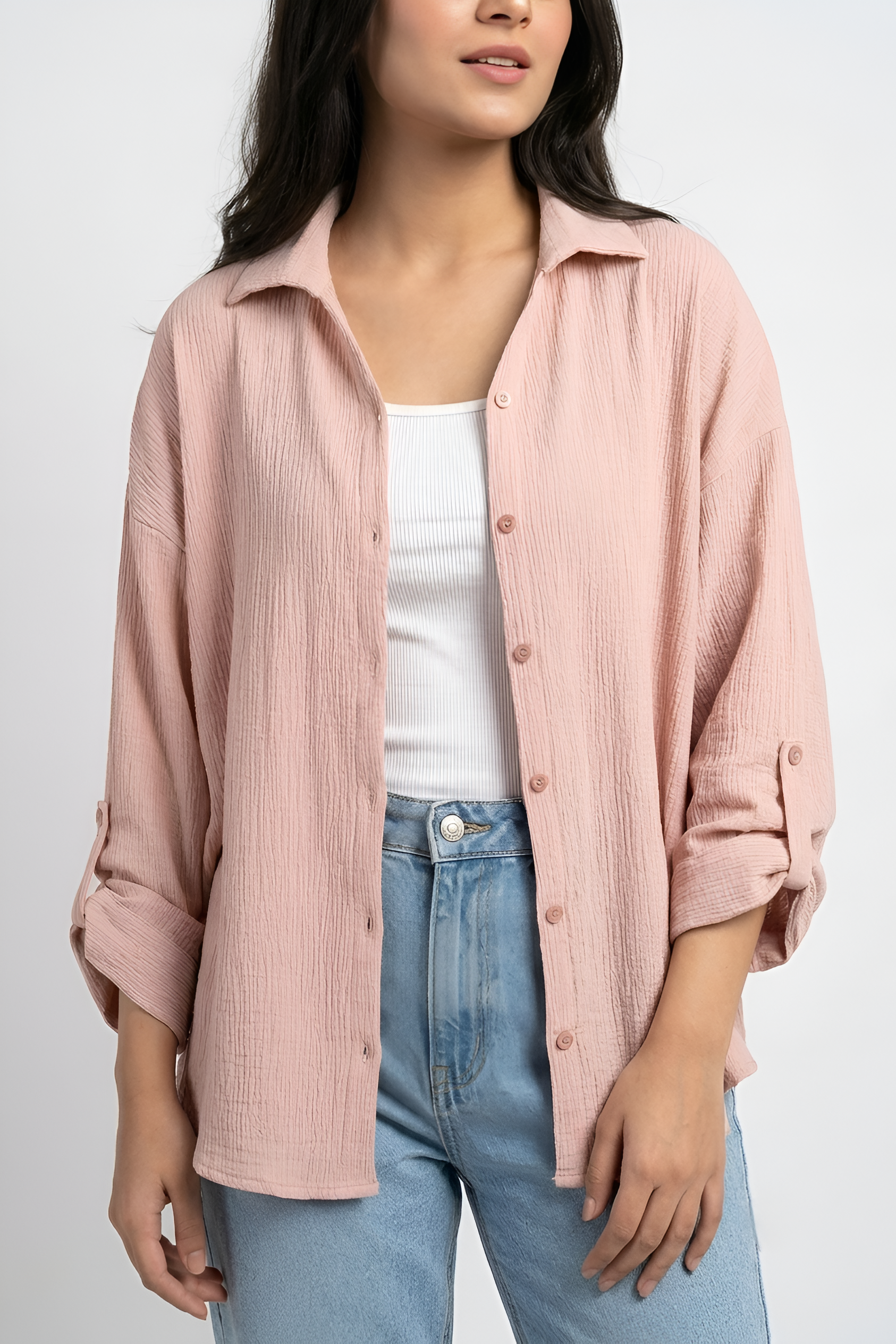 A woman wearing a light pink crinkled cotton button-up shirt over a white tank top and light blue jeans, photographed against a plain light gray studio background.