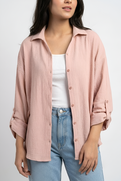A woman wearing a light pink crinkled cotton button-up shirt over a white tank top and light blue jeans, photographed against a plain light gray studio background.