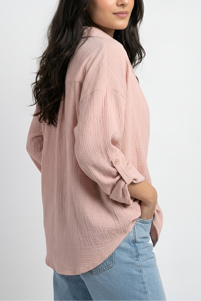 A woman wearing a dusty pink textured long-sleeve blouse and light blue jeans, photographed from the side against a plain light gray studio background.