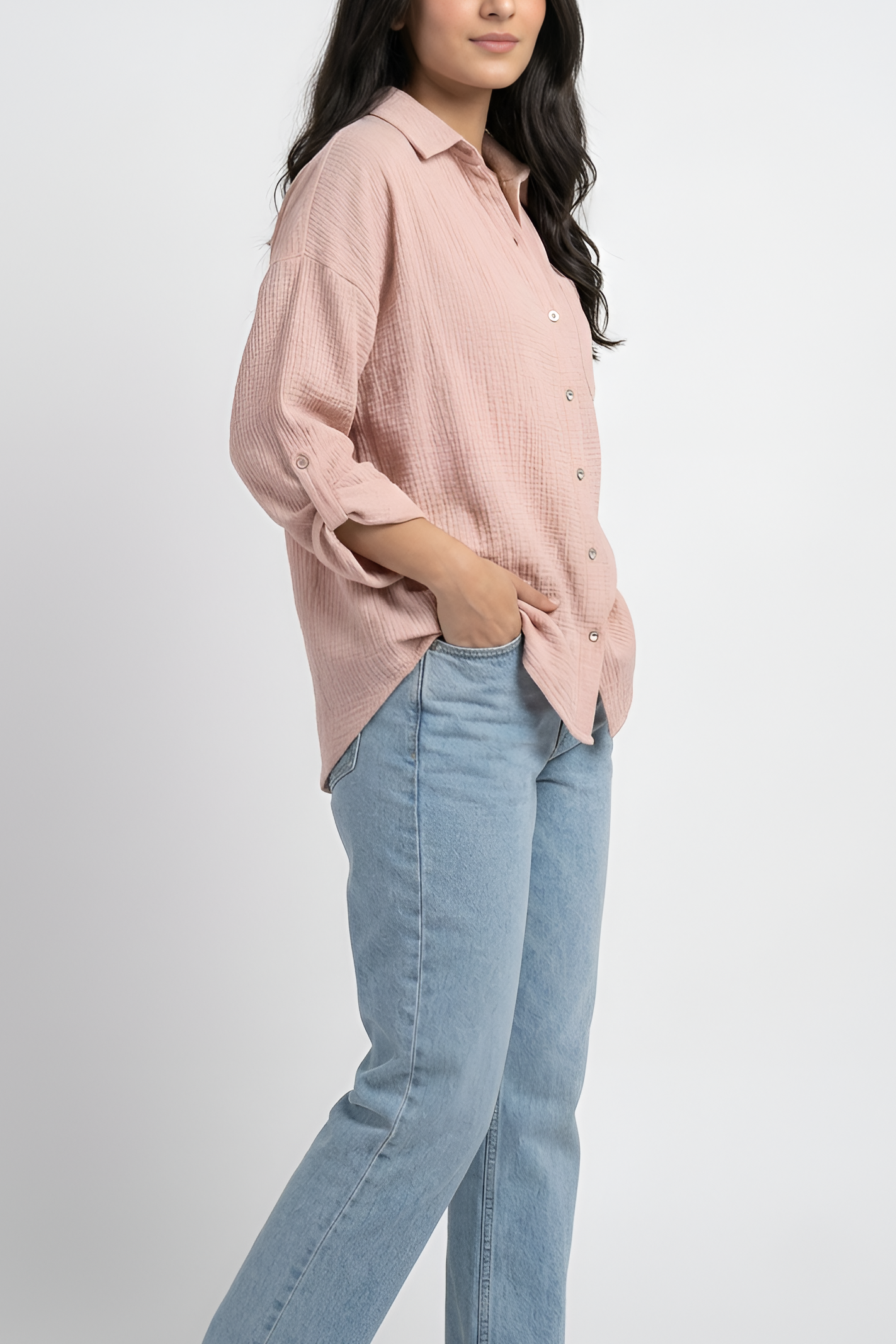 A woman modeling a light pink button-up shirt with light blue jeans, standing against a plain light gray studio background.