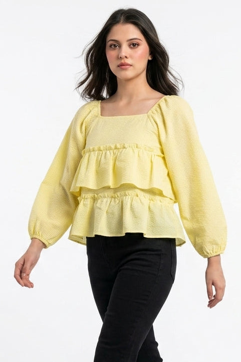 A woman modeling a pale yellow ruffled long-sleeve blouse with a square neckline, paired with black pants and nude high heels, against a plain white studio background.