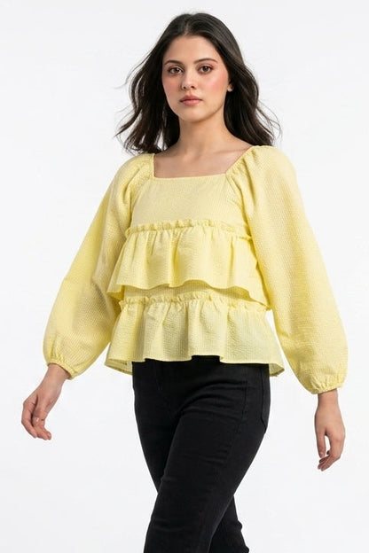 A woman modeling a pale yellow ruffled long-sleeve blouse with a square neckline, paired with black pants and nude high heels, against a plain white studio background.