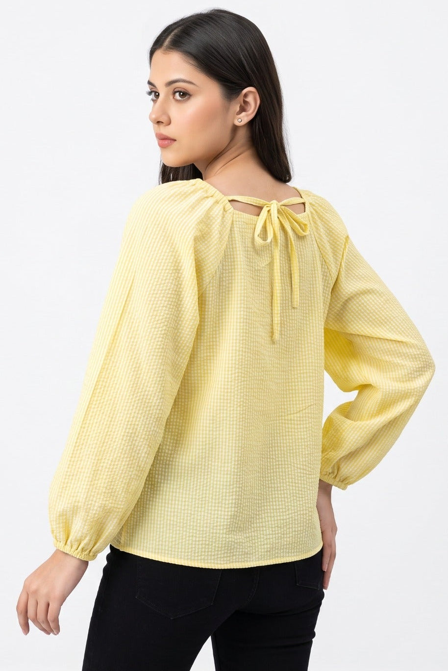 A woman modeling a pale yellow textured long-sleeve blouse with a tie-back neckline, paired with black pants, against a plain white studio background.