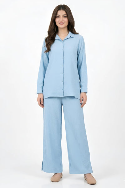 A woman modeling a light blue two-piece pajama set consisting of a long-sleeve button-up shirt and matching wide-leg pants, photographed against a plain white studio background.
