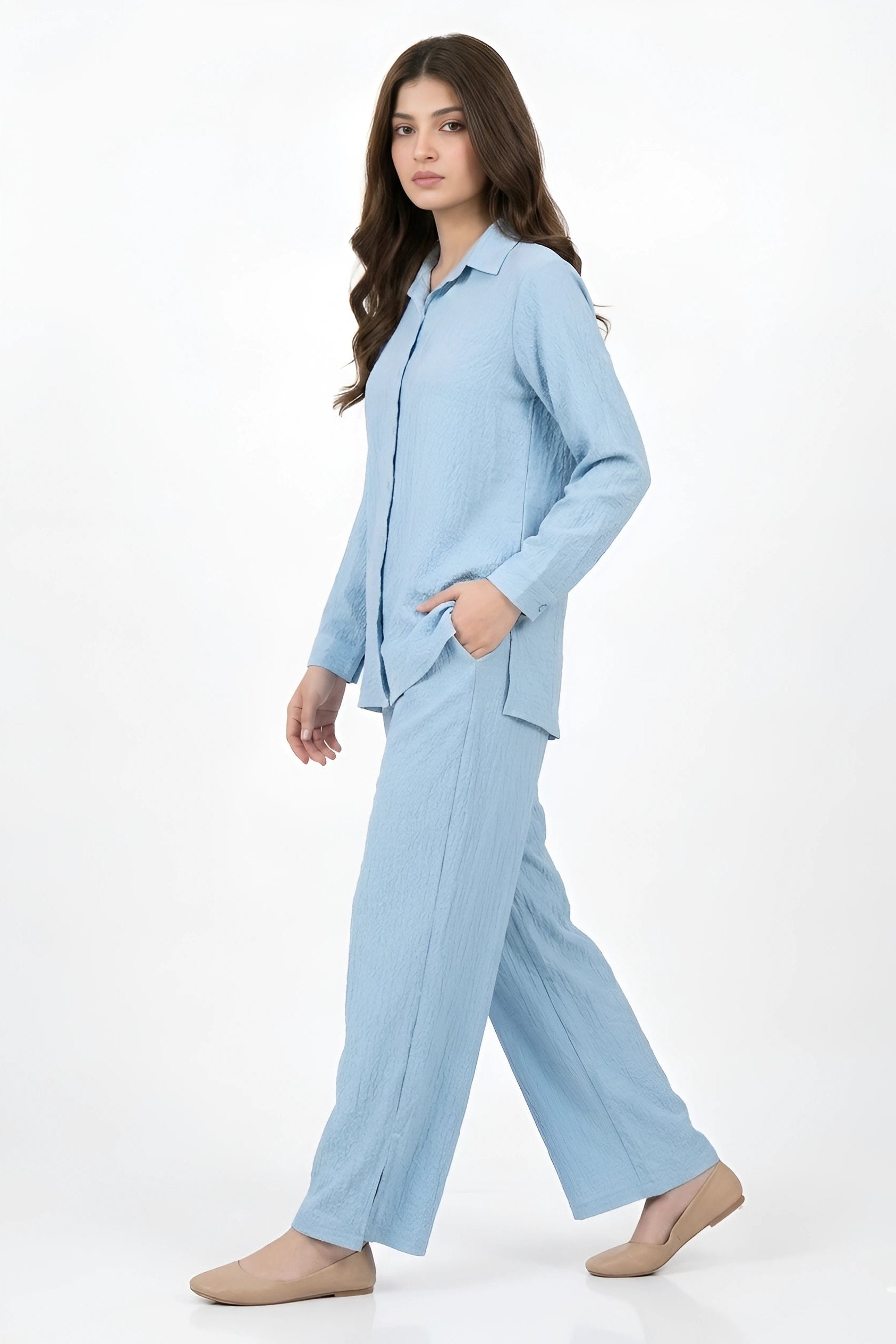 A woman modeling a light blue two-piece pajama set consisting of a long-sleeve button-up shirt and matching wide-leg pants, photographed against a clean white studio background.