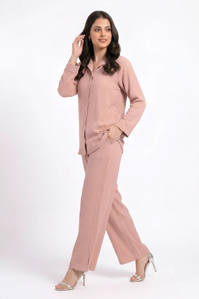 A woman modeling a dusty pink two-piece set consisting of a long-sleeve button-up shirt and matching wide-leg trousers, paired with metallic high-heeled sandals against a plain white background.