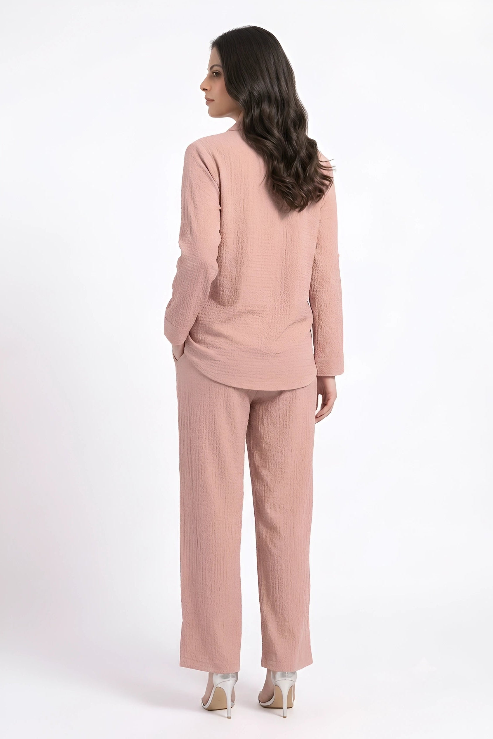 A woman shown from the back wearing a matching dusty pink two-piece set consisting of a long-sleeve cropped jacket and wide-leg trousers, paired with beige high heels against a plain white studio background.