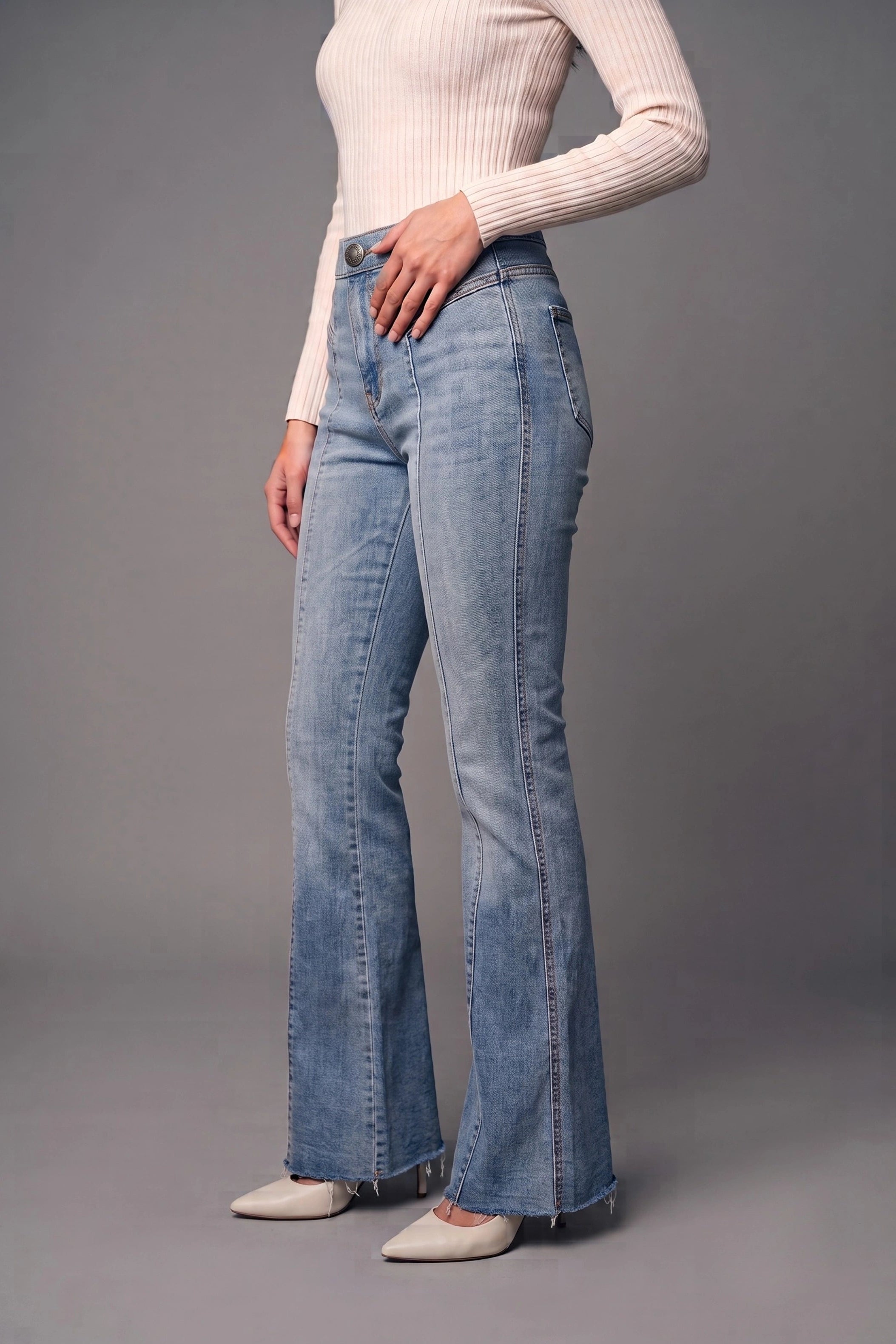 A model wearing light blue high-waisted flare jeans, a cream ribbed long-sleeve top, and beige pointed-toe flats, standing against a solid gray studio background.