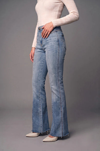A model wearing light blue high-waisted flare jeans, a cream ribbed long-sleeve top, and beige pointed-toe flats, standing against a solid gray studio background.
