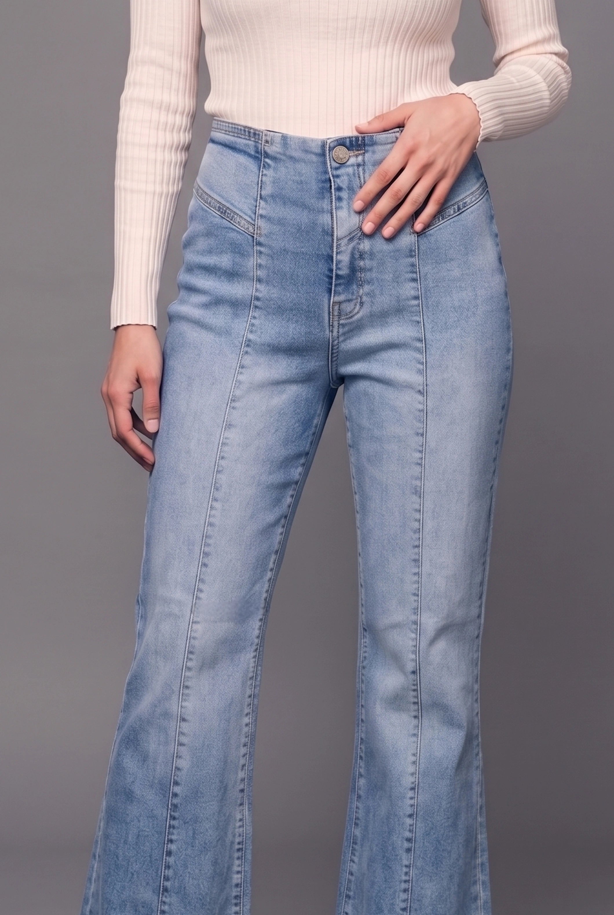 A model wearing light blue high-waisted flare jeans with a cream ribbed knit top, shown from the chest down against a solid gray studio background.