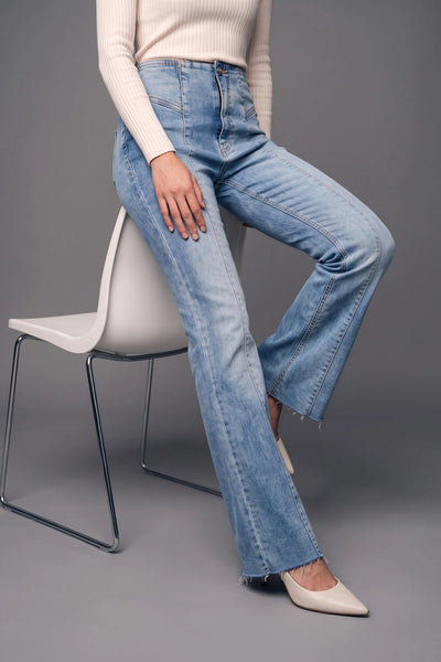 A model wearing light-wash blue flare jeans, a cream ribbed long-sleeve top, and beige pointed-toe flats, seated on a white modern chair against a gray studio background.