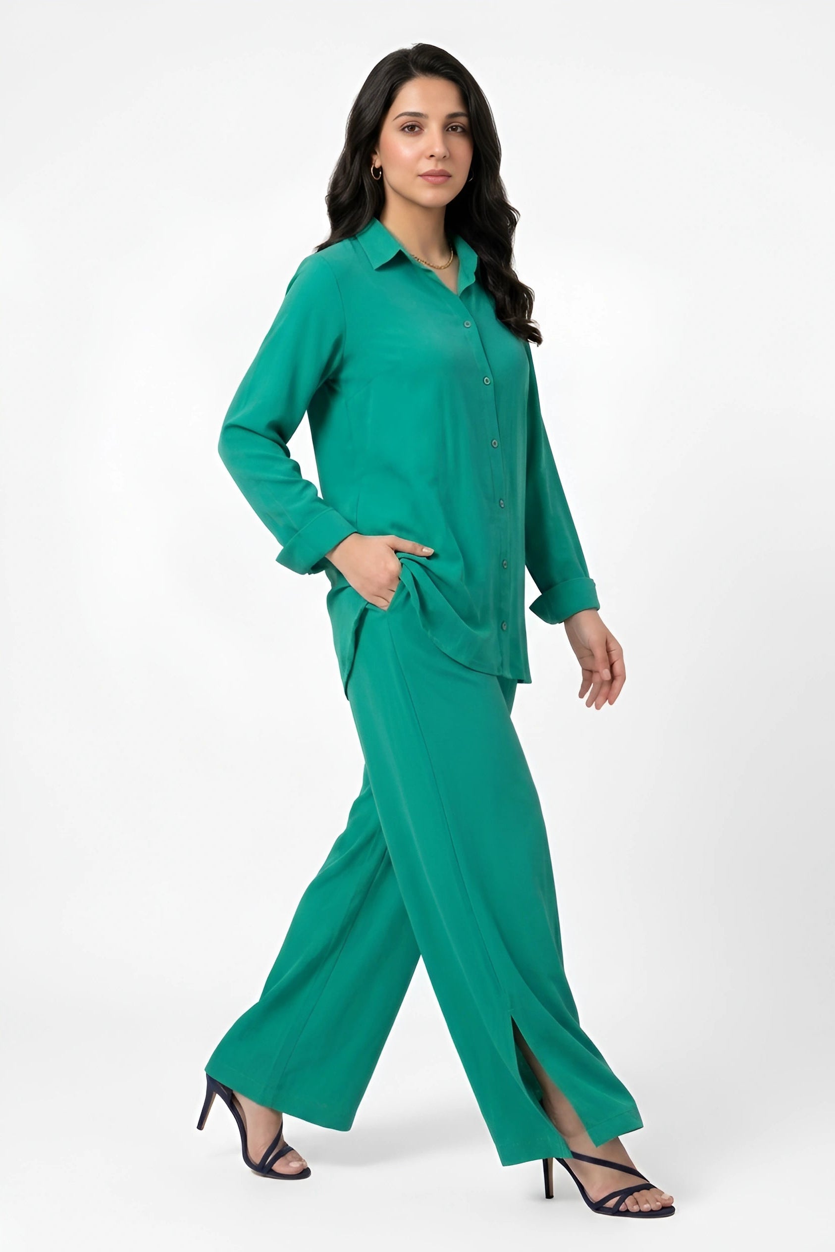 A woman modeling a matching green two-piece set consisting of a long-sleeve button-up shirt and wide-leg trousers, paired with black strappy high-heeled sandals against a plain white studio background.