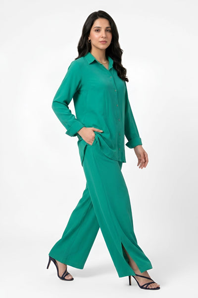 A woman modeling a matching green two-piece set consisting of a long-sleeve button-up shirt and wide-leg trousers, paired with black strappy high-heeled sandals against a plain white studio background.