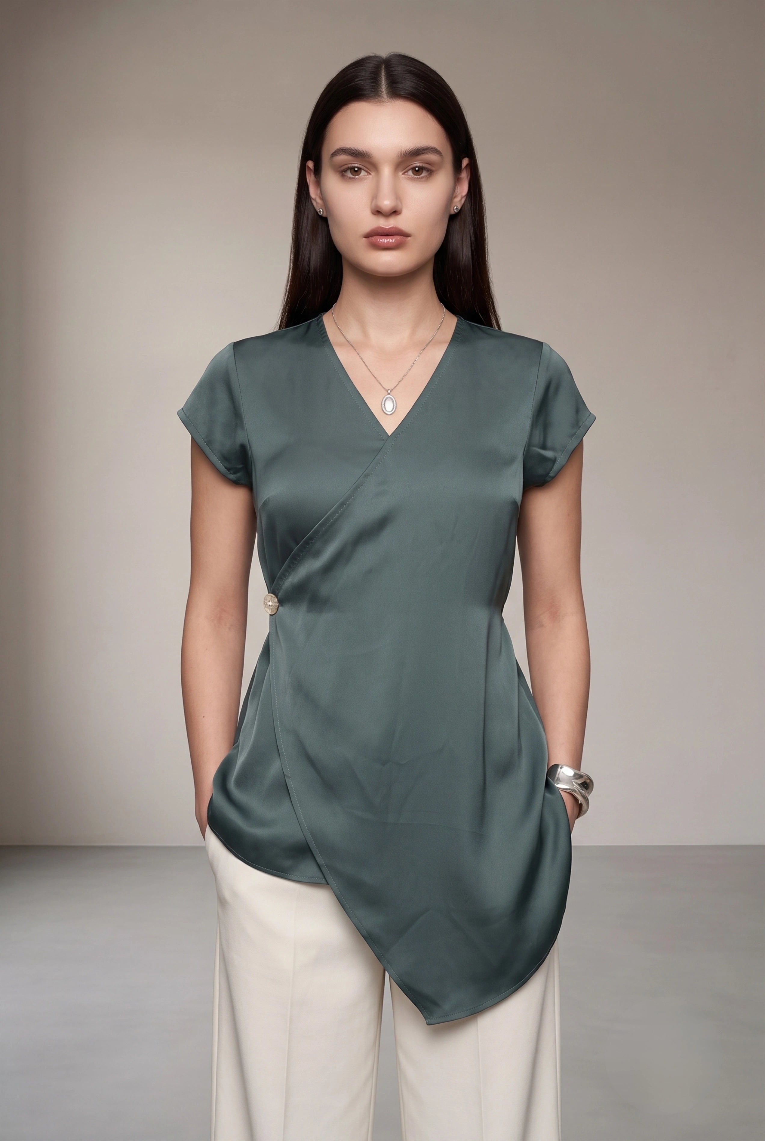 A woman modeling a sage green wrap-style top with short cap sleeves and an asymmetrical draped hem, paired with cream trousers, against a neutral studio backdrop.