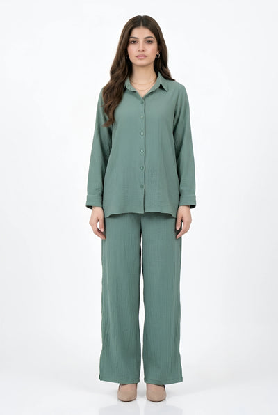 A woman modeling a matching sage green two-piece set consisting of a long-sleeve button-up shirt and wide-leg trousers, photographed against a plain white studio background.