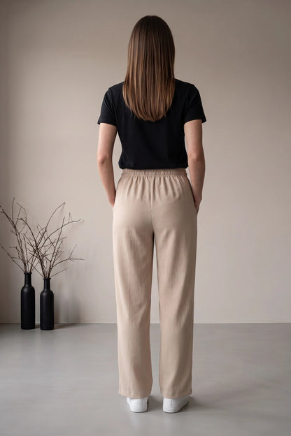 A woman stands with her back to the camera wearing beige wide-leg trousers and a black short-sleeve top, photographed in a minimalist studio setting with two black decorative bottles on the floor.