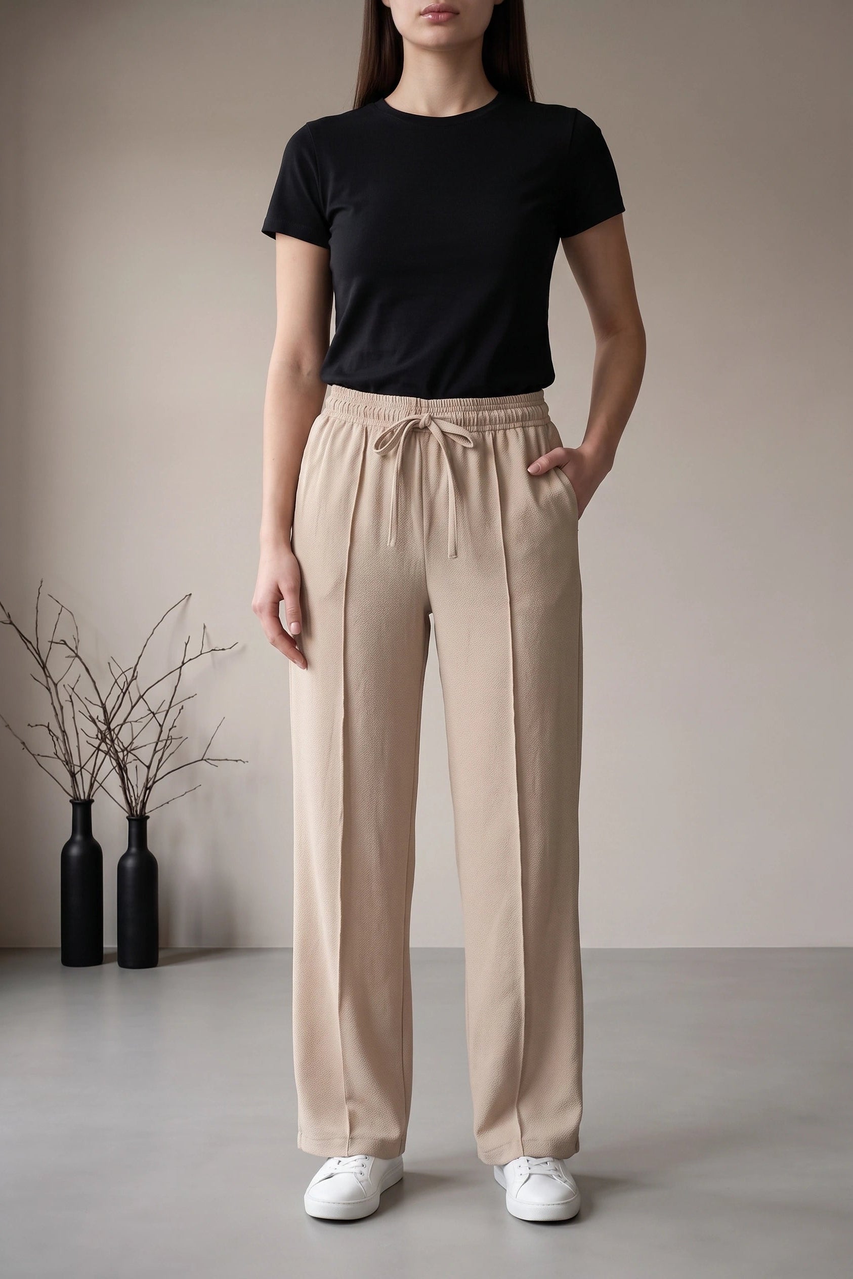 A model wearing beige wide-leg drawstring trousers, a black short-sleeve t-shirt, and white sneakers, standing in a minimalist studio setting with two black decorative bottles on the left.