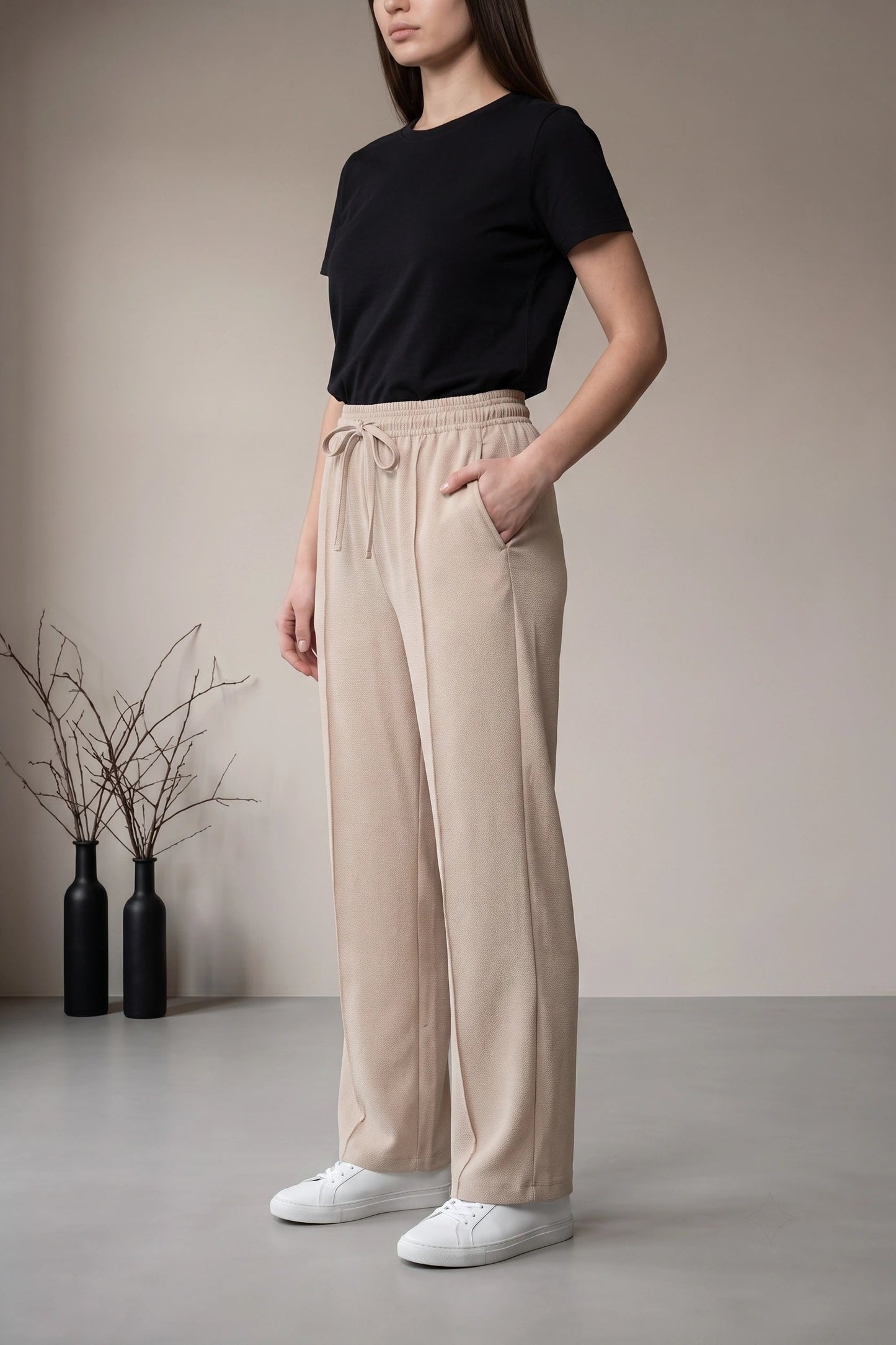 A woman modeling a pair of beige wide-leg drawstring pants with a black t-shirt and white sneakers, standing in a minimalist studio setting.