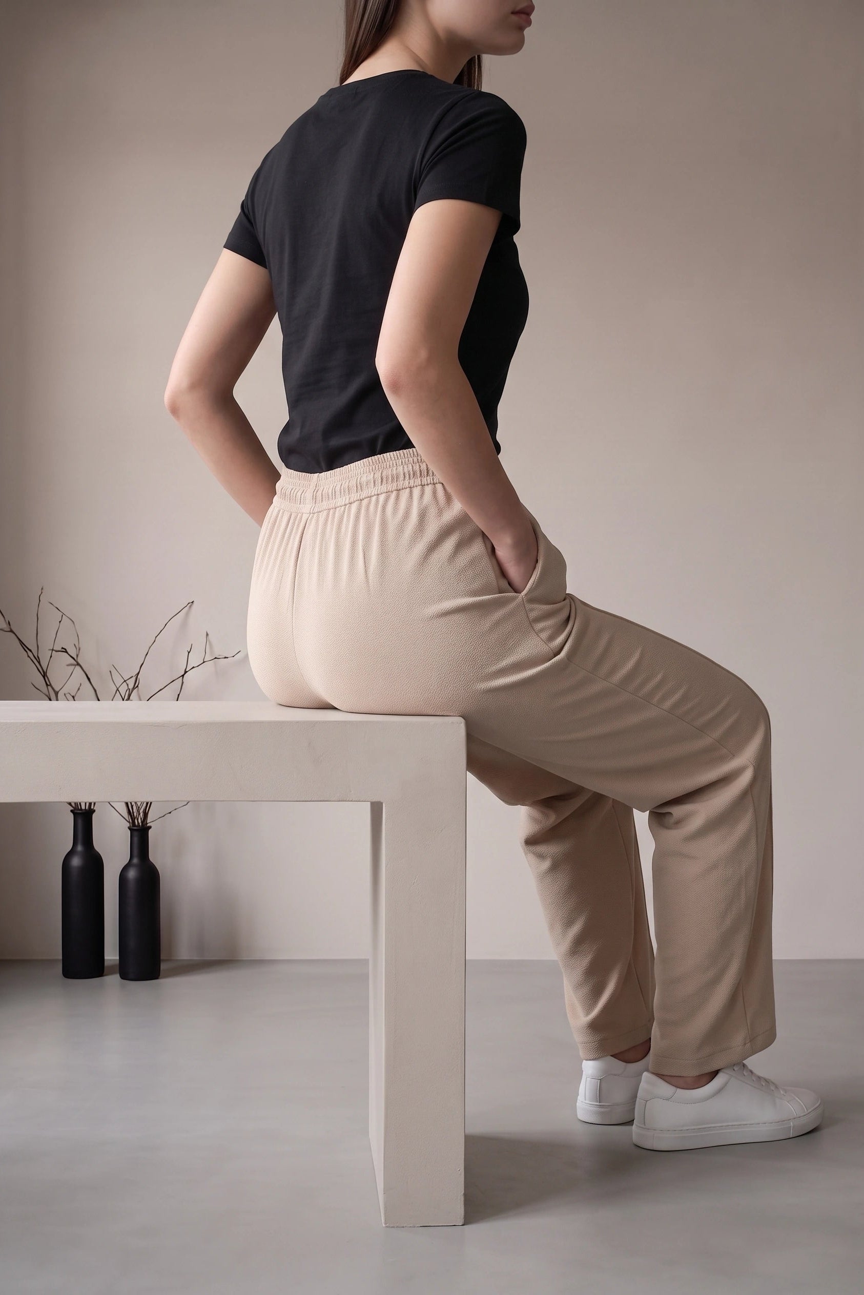A woman seated on a white bench, shown from the back and side, wearing beige high-waisted lounge pants with a black short-sleeve top and white sneakers in a minimalist indoor setting.