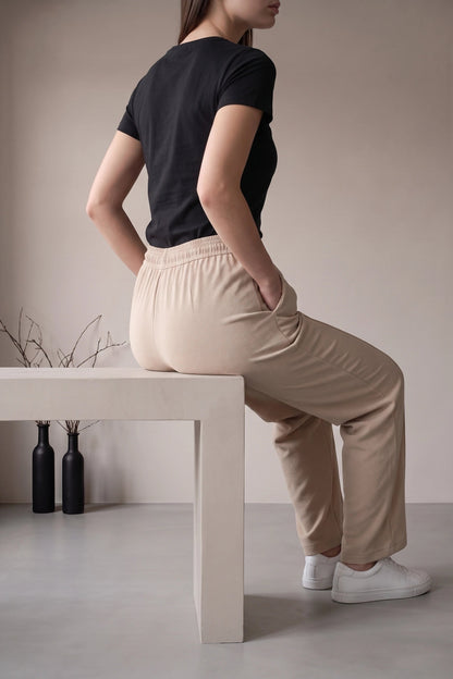 A woman seated on a white bench, shown from the back and side, wearing beige high-waisted lounge pants with a black short-sleeve top and white sneakers in a minimalist indoor setting.