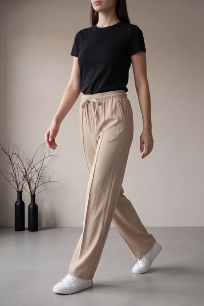 A woman wearing beige drawstring wide-leg trousers, a black short-sleeve t-shirt, and white sneakers, standing in a minimalist studio setting with two black vases containing dried branches.