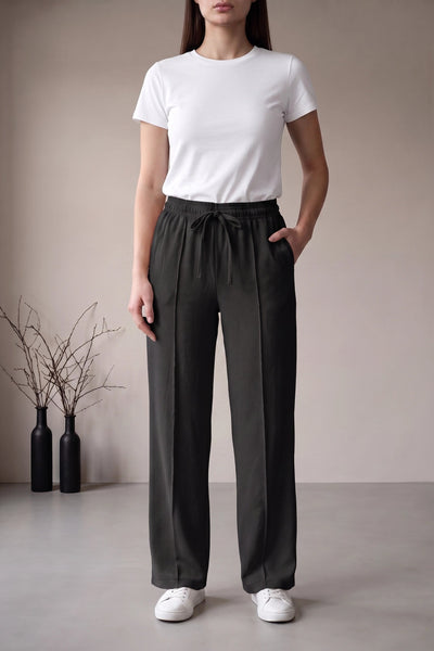 A woman modeling high-waisted dark charcoal gray drawstring trousers with a white t-shirt and white sneakers, standing in a minimalist studio setting.