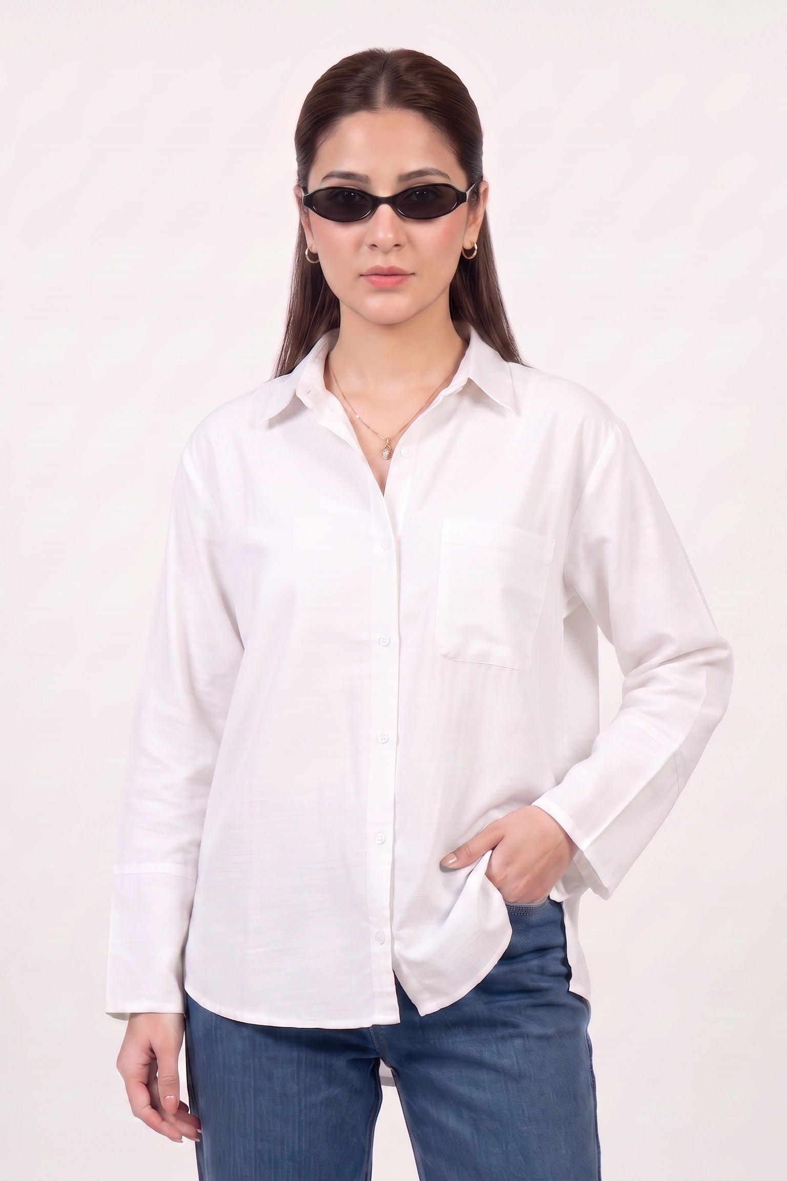 A woman modeling a white long-sleeve button-up shirt with a chest pocket, paired with blue jeans and black sunglasses, against a plain off-white studio background.