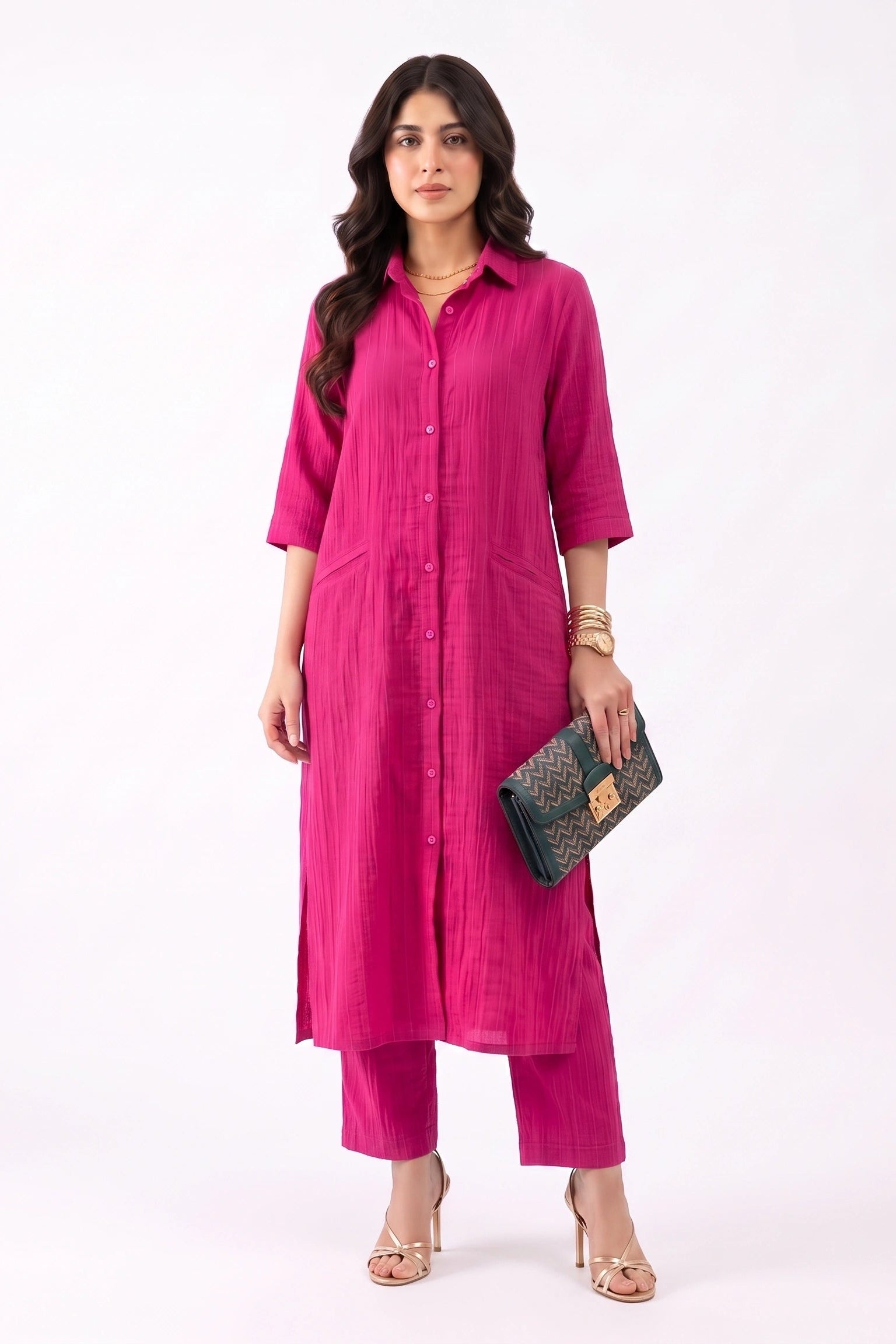 A woman modeling a bright magenta pink kurta set with matching pants, holding a dark patterned clutch, against a plain white studio background.